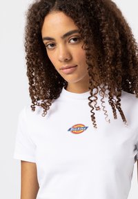 Valge puuvillane T-särk, millel on rinnal ümmargune sinine ja oranž "Dickies" logo. Lühikesed varrukad ja keha lähedane disain.