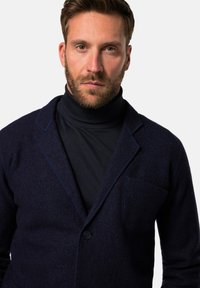 Marineblaue Wollmischjacke mit Schalkragen, einem einzelnen Knopf und aufgesetzter Tasche. Getragen über einem dunklen Rollkragenpullover mit strukturiertem Finish.