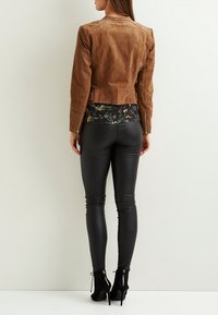 Chaqueta de ante marrón con una forma ajustada, combinada con leggings de cuero negro brillante. El conjunto presenta patrones florales intrincados en la parte superior.