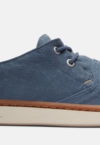 Zapatilla de lona azul con perfil bajo, punta redonda y suela de goma. Presenta ojales de metal y cordones, con costuras en contraste a lo largo de los bordes.