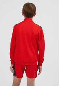 Red athletic tracksuit featuring a long-sleeve jacket and shorts, made of textured fabric with black accents on the shoulders and sides.