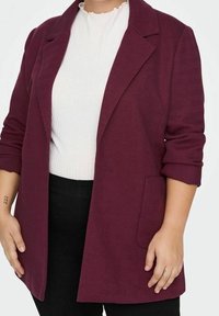 Blazer bourgogne avec une finition texturée, revers crantés et poches avant, porté sur un haut blanc à côtes. Un pantalon noir est visible.