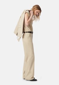 Beige op maat gemaakt pak met een nauwsluitend bovenstuk en broek met wijde pijpen. Het model draagt een bijpassend colbert over haar schouder. Zwarte riem en schoenen.