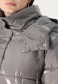 Jakna s punjenjem u metalno sivoj boji, s visokim ovratnikom, šivenim panelima i kopčama s dugmetima. Glatka tekstura s suptilnim sjajem i naglascima.