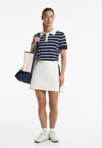 Navy and white striped polo shirt with a collar, paired with a white skort featuring side zippers; model holds a blue and cream tote bag.
