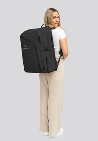 Sac à dos noir avec plusieurs zips et boucles, arborant un logo brodé. Texture lisse, design structuré et bretelles ajustables.