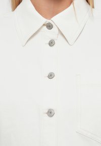 Valge kraega särk, millel on viis hõbedast nuppu, rinnatasku ning sile tekstuur peente õmblusdetailidega.