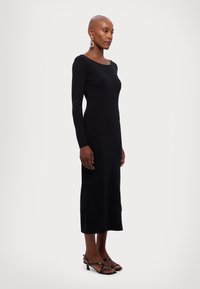 Abito lungo in maglia nera a maniche lunghe con scollatura quadrata, silhouette aderente e lunghezza fino al polpaccio. Abbinato a sandali neri con tacco e fasce.