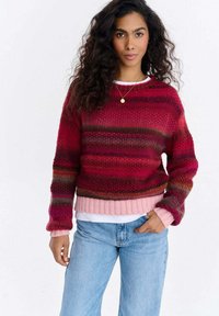 Sweter na drutach w odcieniach czerwieni z delikatnym gradientem. Długie rękawy, ściągacze na mankietach i u dołu oraz warstwowo nałożony na białą koszulę. Miękka tekstura.