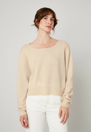 Pull en tricot beige avec un grand col rond et des manches longues, doté d'un ourlet côtelé et d'une finition douce et texturée. Porté avec un jean blanc.