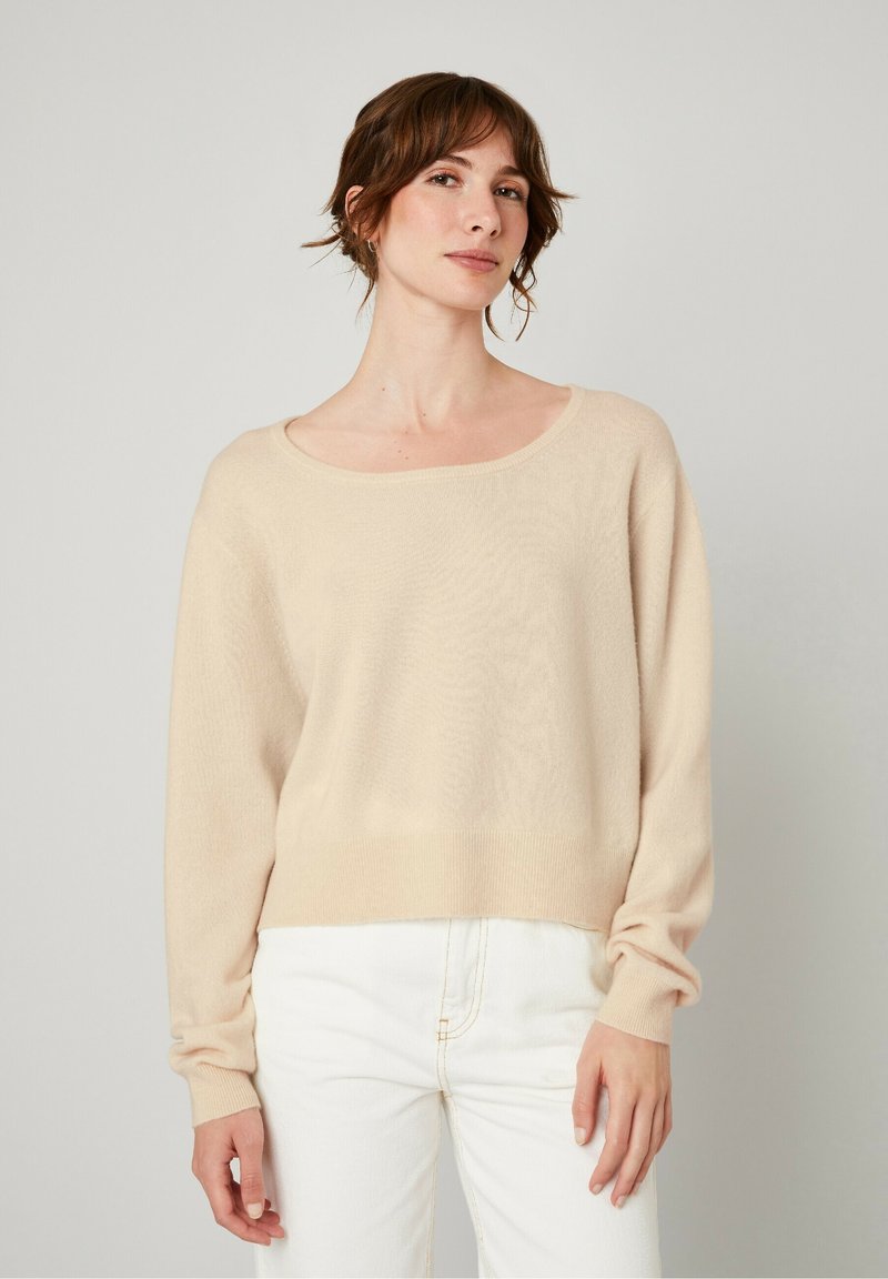 Pull en tricot beige avec un grand col rond et des manches longues, doté d'un ourlet côtelé et d'une finition douce et texturée. Porté avec un jean blanc.