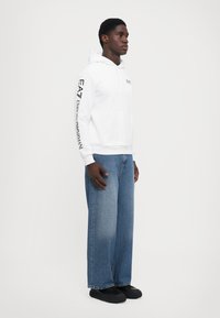 Sweatshirt branca com capuz e logótipo na manga, bolso frontal, combinado com jeans azuis de ajuste solto e sapatos pretos. Design minimalista.