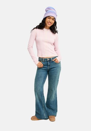 Frau lächelt mit den Händen in den Taschen, trägt ein pinkes Langarmoberteil, blaue ausgestellte Jeans, braune Schuhe und eine lila gestrickte Mütze.