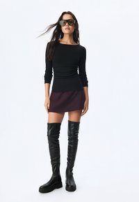 Schwarzes Langarmoberteil kombiniert mit einem bordeauxroten Rock, ergänzt durch kniehohe schwarze Lederstiefel. Das Modell trägt große schwarze Sonnenbrillen.