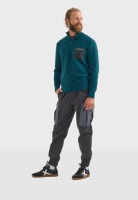 Pullover en polaire bleu canard avec fermeture éclair half-zip et une poche poitrine noire, associé à un pantalon cargo noir avec poches grises et poignets élastiques. Baskets noires.