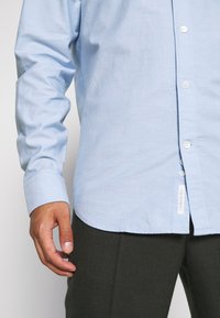 Homme portant une chemise bleu clair à manches longues boutonnée et un pantalon sombre, debout avec la main gauche relâchée sur le côté.