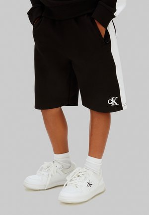 Bambino che indossa pantaloncini neri con strisce laterali bianche e sneakers bianche, entrambe con il logo cK, in piedi con le mani nelle tasche.