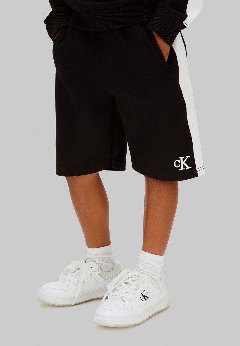 Niño con pantalones cortos negros con rayas blancas a los lados y zapatillas blancas, ambos con logos de cK, de pie con las manos en los bolsillos.