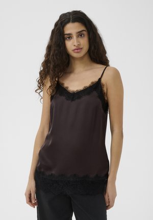 Donna con capelli lunghi e ricci che indossa un top in satin nero con bordi in pizzo, in piedi su uno sfondo chiaro e uniforme.