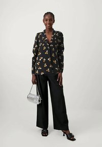 Sieviete, tērpta melnā ziedu blūzē, platās melnās biksēs, melnos sandalos ar papēžiem, tur nelielu sudraba rokassomu ar ķēdes siksnu.