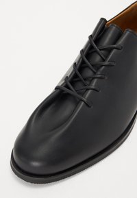 Black leather shoe featuring a rounded toe, sleek design, and lace-up detail. Smooth texture with a subtle shine, low profile rubber sole.