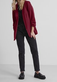 Blazer rouge à manches retroussées sur un haut noir, assorti à un jean noir taille haute et des mocassins noirs épais. Textures lisses et coupe ajustée.