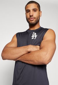 Top atlético sin mangas de color gris oscuro con una textura suave, que presenta un logo blanco "LA" en el lado izquierdo del pecho. El diseño enfatiza la comodidad y la movilidad.