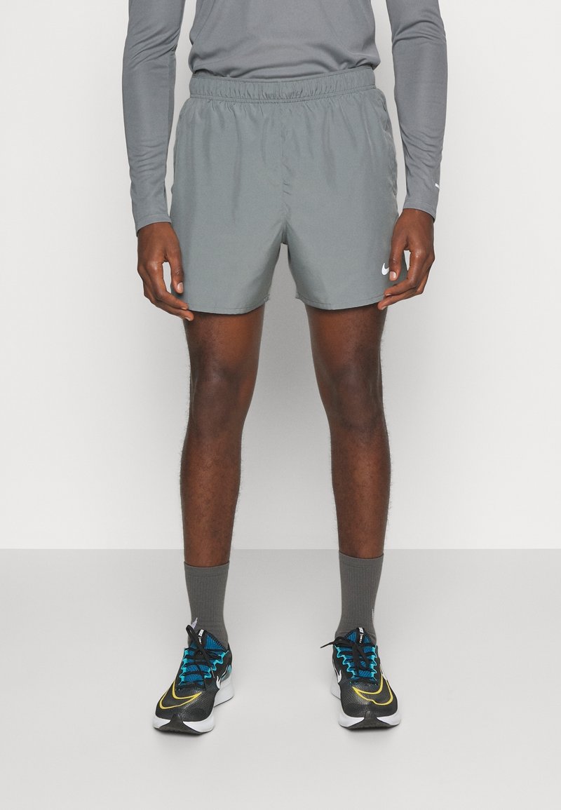 Homme portant un short de sport gris, un haut à manches longues gris, des chaussettes grises et des chaussures de course noires avec des accents bleus et jaunes, debout sur fond blanc.
