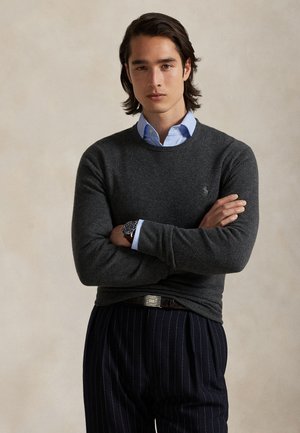 Jeune homme aux cheveux mi-longs foncés portant un pull gris sur une chemise bleue, un pantalon sombre rayé et une montre-bracelet, debout les bras croisés.