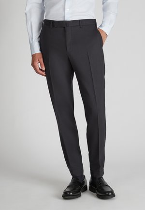 Maßgeschneiderte dunkelgraue Hose mit flachem Frontschnitt, ausgestattet mit Gürtelschlaufen und einem Bügel auf den Beinen. Getragen mit schwarzen Slippern.