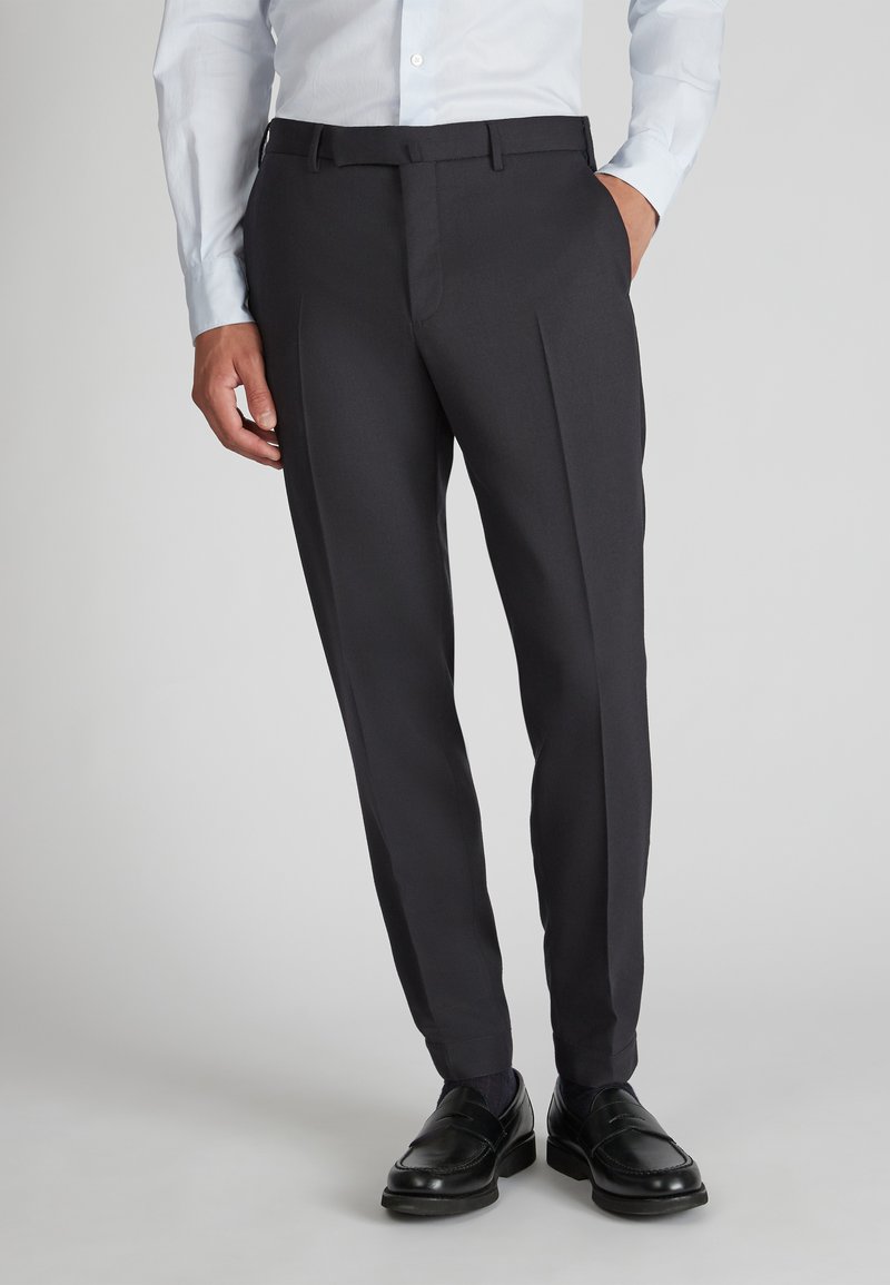 Maatwerk donkergrijze broek met een vlakke voorkant, voorzien van ceintuurlussen en een plooi langs de benen. G gedragen met zwarte instappers.