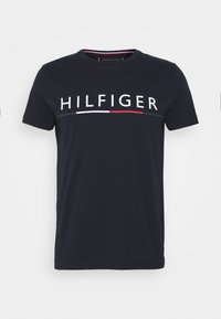 Granatowa bawełniana koszulka z okrągłym dekoltem, krótkimi rękawami i wyrazistą białą grafiką "HILFIGER" z czerwonymi i białymi paskami poniżej.