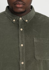 Camicia in corduroy verde con colletto button-down, cinque bottoni chiari e taschino sul lato sinistro del petto. Tessuto testurizzato con righe verticali.
