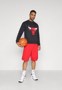 Melnā sporta krekls ar sarkano bulli grafiku, sarkanas šortus, baltas Nike kedas un turot basketbola bumbu. Vienkāršs dizains, viegla auduma.