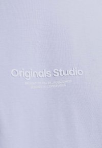 Baltos marškinėliai su reljefiniu tekstu, kuriame parašyta "Originals Studio", ir papildomi prekės ženklų elementai šiek tiek tamsesnėje spalvoje.