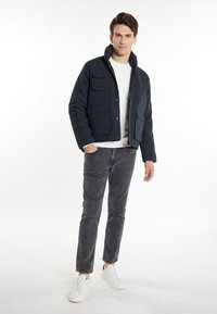 Navyblauer Puffer-Jacke mit vier Fronttaschen, einem Stehkragen und einem Reißverschluss, kombiniert mit grauen Hosen und weißen Sneakers.