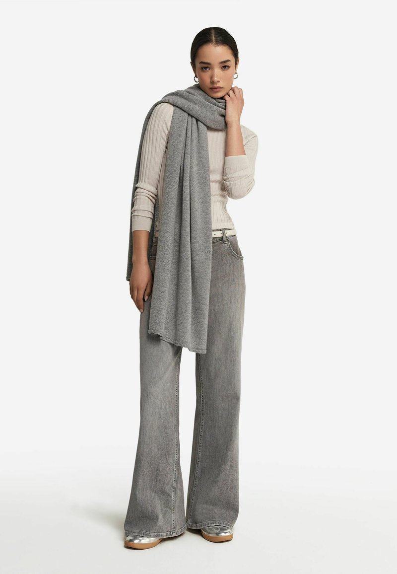 Une écharpe en laine grise drape un haut beige clair à côtes et un jean flare gris taille haute, complété par des chaussures argentées et une ceinture blanche.