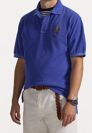 Mężczyzna w niebieskiej koszulce polo z małą naszywką niedźwiedzia, białych spodniach z plecionym brązowym paskiem oraz zegarku na nadgarstek z niebieską tarczą i brązowym skórzanym paskiem.