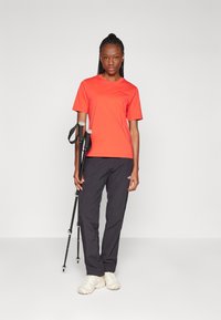 T-shirt rouge à manches courtes, pantalons noirs et chaussures claires. La personne tient des bâtons de trekking, et la tenue est conçue pour l'activité de plein air.