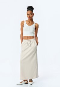 Top court blanc à côtes avec détails de boutons associé à une longue jupe beige avec taille élastique et poches. Chaussures blanches avec brides.