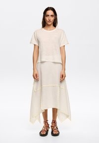 Cream linen top with short sleeves, paired with a flowing cream skirt featuring a scalloped hem and yellow accents. Black sandals with red straps.