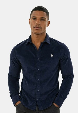 Junger Mann mit kurzen Haaren trägt ein navyblaues, geknöpftes Langarmhemd mit kleinem weißem besticktem Logo auf der Brust.