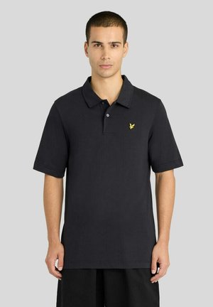 Ung mand med kort hår iført en sort polo-shirt med en gul fuglelogo, stående med arme afslappet mod en grå baggrund.