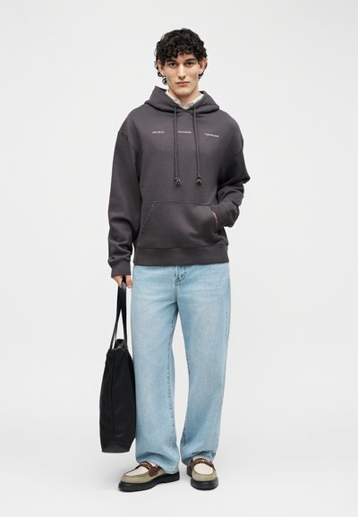 Jaunas vyras su garbanotais plaukais dėvi tamsiai pilką džemperį su gaubtu, šviesiai mėlynas džinsus ir dviejų spalvų batus, laiko juodą nešiojamą krepšį priešais paprastą foną.