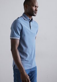 Cămașă polo albastră deschisă cu guler cu două nasturi, guler contrastant și dungi pe manșetele mânecilor. Fabricată dintr-un material neted, are un design ajustat.