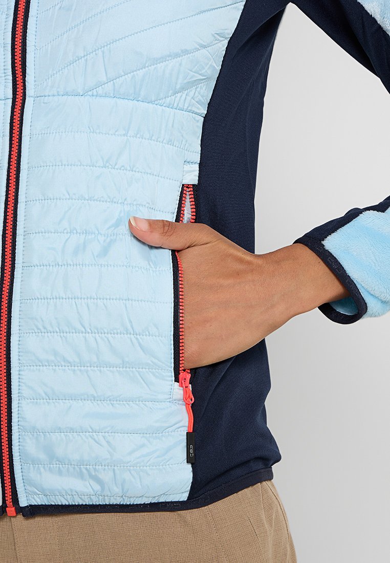 Veste rembourrée bleu clair avec un motif matelassé horizontal, manches navy et accents de fermeture éclair rouge vif. Un main repose sur une poche latérale.
