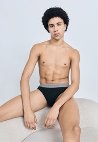 Fekete alsónadrág szürke derékrésszel, rajta a "SCHIESSER" márkajelzés. A modell egy világos, texturált felületen ül, semleges háttér előtt.