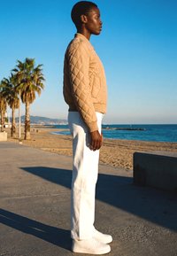 Person in beige gesteppter Jacke und weißer Hose steht auf einer Strandpromenade neben Palmen unter klarem blauem Himmel.