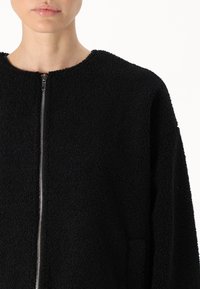 Giacca in fleece nera con scollatura tonda e zip frontale. Il tessuto ha una superficie texturizzata e morbida. Presenta spalle scese e tasche laterali.