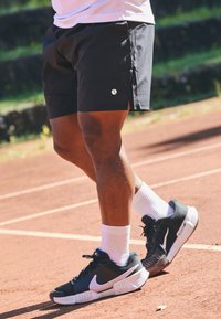 Calções desportivos pretos com fendas laterais, combinados com ténis Nike preto e branco e meias brancas, num campo de ténis de argila.